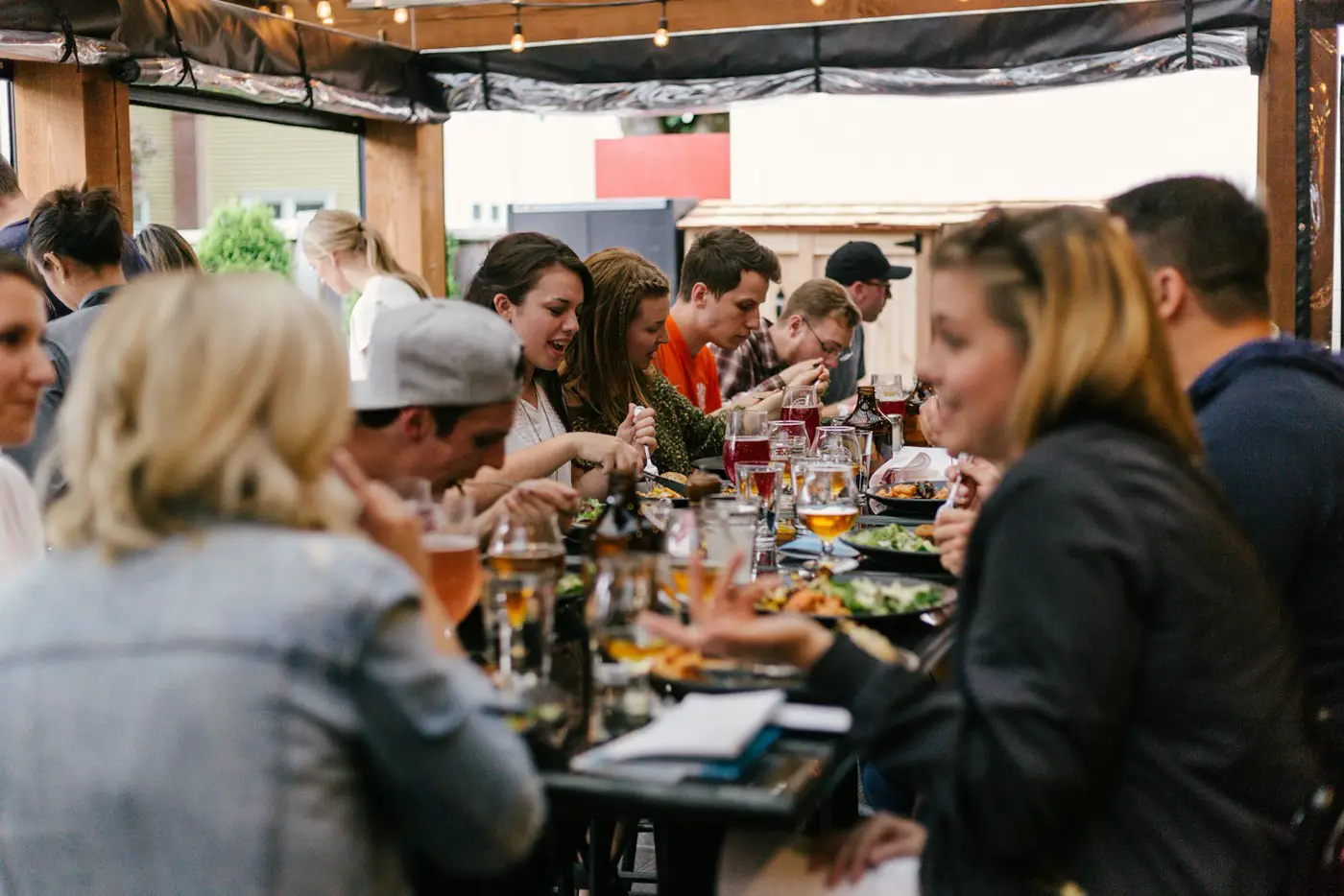 A long outdoor table on a busy terrace, friends leaning in to talk over food and drinks