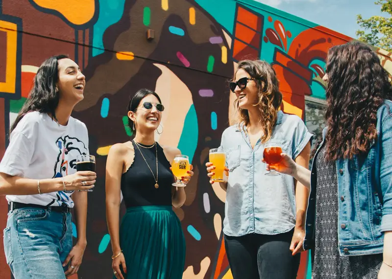 Four friends laughing in front of a colourful painted mural on a sunny street, drinks in hand